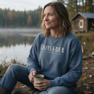 Woman wearing a faded blue Outsider unisex crewneck sweatshirt while enjoying coffee on a wooded cabin porch