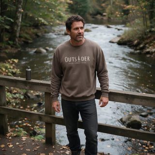 Man wearing an espresso Outsider crewneck sweatshirt near a wooded river trail