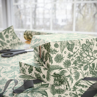 Stack of presents wrapped in green and cream evergreen toile wrapping paper by a window