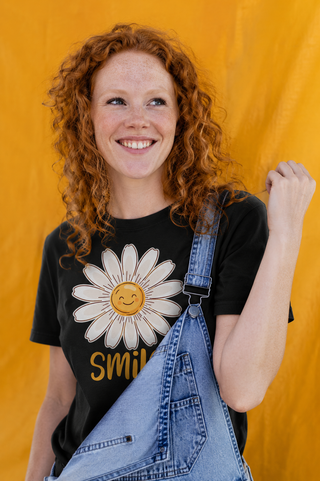 Woman wearing the Artisan Grove Sunshine Daisy Smile T-shirt with a cheerful hand-drawn daisy graphic, styled with denim overalls in a bright outdoor setting.