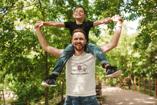 Man carrying a child on his shoulders with a 'Farmer Market' t-shirt in a park setting