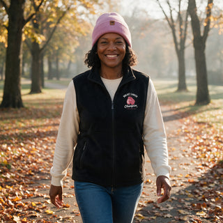 Smiling woman on a fall park walk in the black Breast Cancer Champion fleece vest and a light pink cuffed beanie