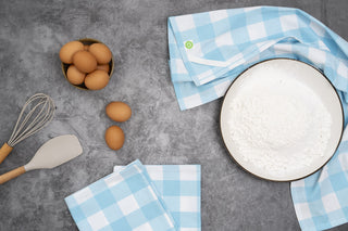 Light blue plaid kitchen towels displayed with flour, eggs, and baking utensils
