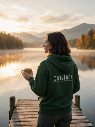 Woman holding a coffee mug on a lake dock at sunrise wearing a forest green Outsider hoodie with Explore Wonder Belong on the back