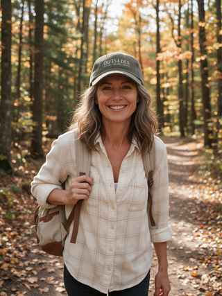 Smiling woman on an autumn forest trail wearing the olive Outsider hat with embroidered pine tree logo and Explore Wonder Belong