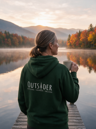 Woman with a ponytail watching a misty autumn lake at sunrise in a forest green Outsider pine tree hoodie